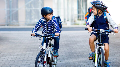 Zwei Kinder fahren lachend nebeneinander auf Fahrrädern. Sie haben Helme und Schulranzen auf.