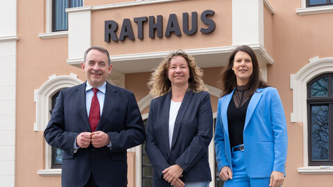 Drei Personen – ein Mann im Anzug und zwei Frauen in Blazern – stehen lächelnd vor dem Eingang eines rötlichen Gebäudes mit dem Schriftzug „RATHAUS“.