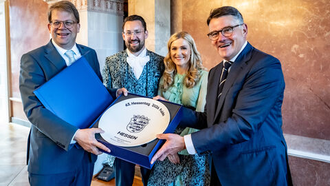 Vier lächelnde Personen präsentieren gemeinsam einen großen, runden Ehrenteller des Hessentages 2026 in Fulda in einer blauen Schachtel.Eine große Konferenzrunde in einem historischen Saal mit zwei prächtigen Kronleuchtern, bei der die Teilnehmer an einer u-förmigen Tischreihe tagen.